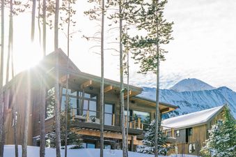 A modern lodge, Moonlight Basin Lodging 7 Rolling Ridge, with large windows and wooden siding, is surrounded by snow-covered pine trees and majestic mountains under a bright winter sky.