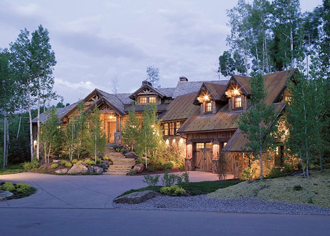 A large, multi-level chalet-style home at dusk, with warm lights glowing from its windows, nestled among tall trees, with a winding driveway and landscaped gardens leading to its entrance.