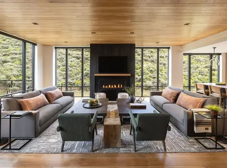 A luxurious living room in The Sylvan Lodge Two Bedroom Penthouse featuring two large gray sofas, a fireplace, and expansive windows overlooking a lush green landscape.