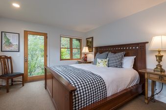 A cozy bedroom with a queen-sized bed, white linens, a black and white buffalo check runner, and two bedside tables with lamps, offering views of lush greenery through a window and a glass-paneled door.