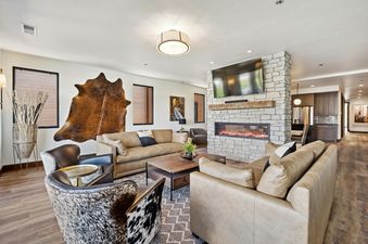 The spacious living area at Moonlight Ridge features tan leather sofas, cowhide accent chairs, a stone fireplace with a mounted television, and rustic-chic decor, providing a warm and inviting space.