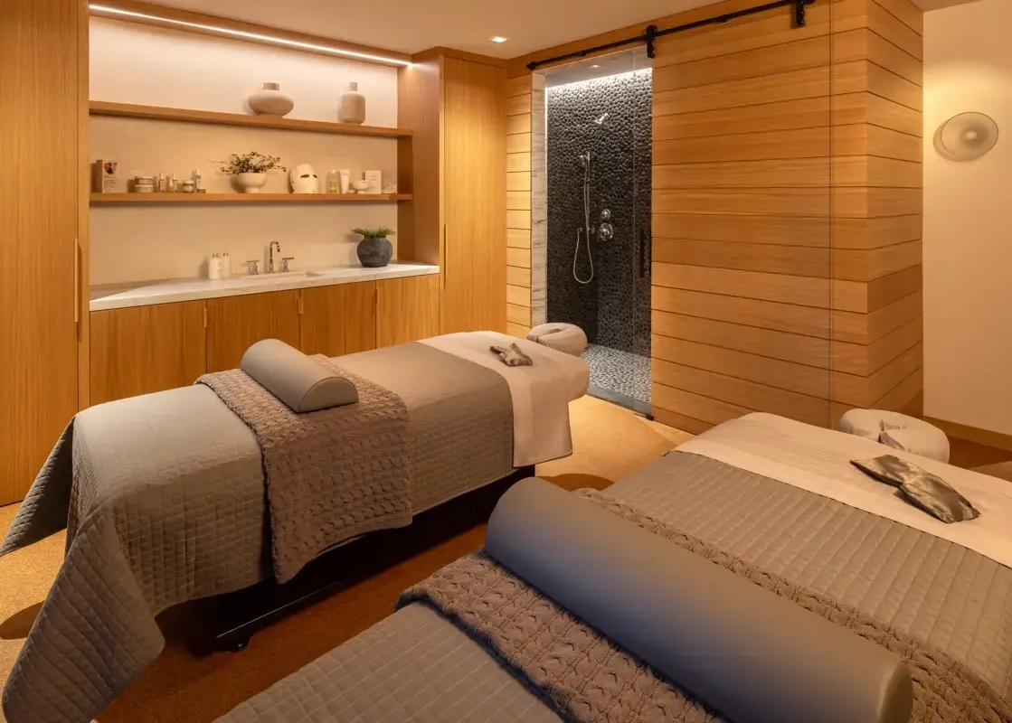 A luxurious massage room in The Sylvan Lodge Two Bedroom Suite features two massage beds with gray linens and a private shower with pebble walls.