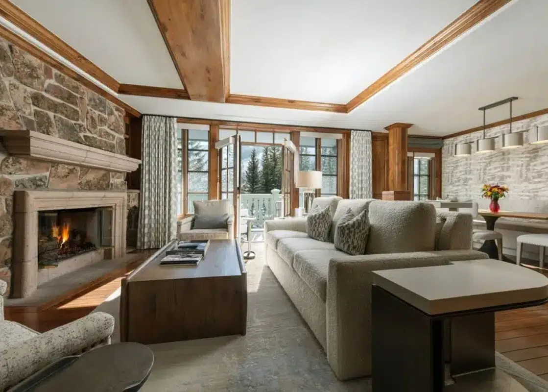 The spacious living room and dining area of Chateau Beaver Creek Residence 204 features a stone fireplace, comfortable sofas, and large windows with a view of snow-covered trees.