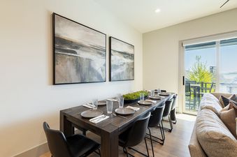 A modern dining area with a long wooden table set for several guests, featuring abstract artwork on the wall and a sliding glass door leading to an outdoor balcony.