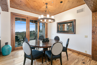 The dining room in Grand Lodge Three Bedroom Deluxe 407 features a large wooden table with six chairs, a chandelier hanging from the ceiling, a mirror on the wall, and large windows overlooking the mountains.