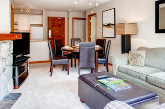 The dining area and living room of Aspens on Blackcomb Unit 547 are showcased, featuring a round wooden dining table with leather chairs and a comfortable sofa.