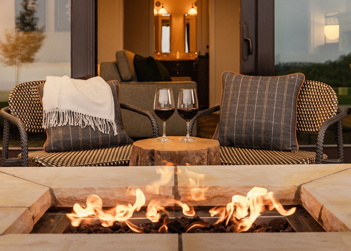 An outdoor patio features two chairs with pillows, a small wooden table, and two glasses of red wine, all centered around a glowing fire pit, with an interior room visible in the background.