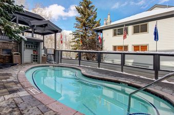 An inviting outdoor hot tub at Snow Flower 152 Park City, surrounded by snow-dusted paving and pine trees under a clear blue sky, with a building featuring flags in the background.