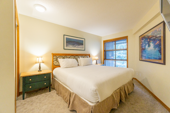 The primary bedroom features a large, comfortable bed with crisp white linens, flanked by matching nightstands with lamps, and a window with blinds.