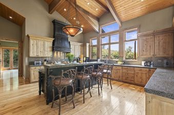 A kitchen with wooden cabinets, granite countertops, and a large island with four bar stools. The kitchen has a vaulted ceiling with exposed wooden beams and large windows overlooking a forested area.