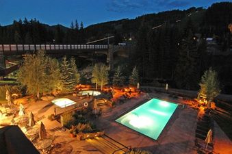 An elevated view of a resort pool and hot tubs lit up at night, with a bridge and forested mountains in the background under a dark blue sky.