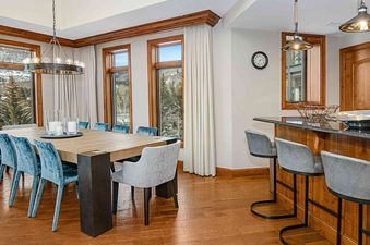 The dining room at Ritz Carlton Vail 402, showcasing a sleek table with chairs, large windows overlooking a snowy mountain landscape, and a chandelier hanging from the ceiling.