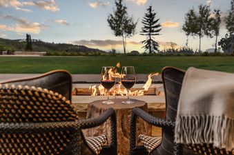 Two chairs face a fire pit with two glasses of red wine on a small wooden table, set against a backdrop of a green lawn, distant trees, and a sunset sky.