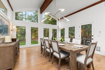 A brightly lit dining area in the Alpine Retreat Jackson Hole with a large wooden table, white upholstered chairs, and tall windows showing forest views.