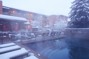 An outdoor pool at the Aspen Square Hotel Deluxe Three Bedroom steams in the winter snow, surrounded by snow-covered lounge chairs, a dining patio, and trees.