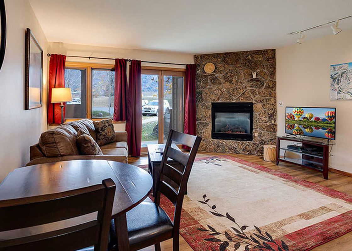 A wide shot of the living and dining area in The Pines Condominiums P103C, featuring a brown sofa, a wooden dining table, a stone fireplace, and a television.