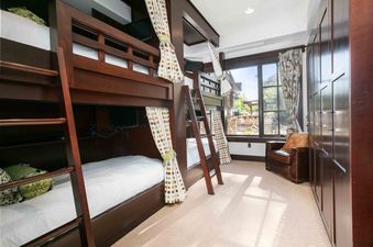 A bunk room with two wooden bunk beds, each with white bedding and privacy curtains, alongside a large window revealing an outdoor patio.