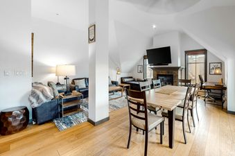 A wide shot of the great room at Main Street Station 3405, featuring an open-concept living area with sofas and a dining area with a wooden table, all with wood flooring and light walls.