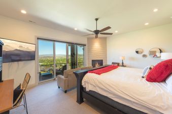 A spacious bedroom in Juniper 801 features a bed with white linens and red accents, a mounted TV, a workspace, a couch, a fireplace, and a ceiling fan, all overlooking a stunning exterior view.