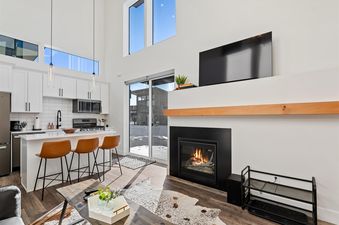 A modern open-concept living area and kitchen with white cabinetry, a kitchen island, leather bar stools, a fireplace, and large windows offering an exterior view.