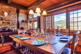 A large dining room at Antler Ridge Lodge features a wooden table set for dinner, a stone fireplace, and large windows with mountain views.