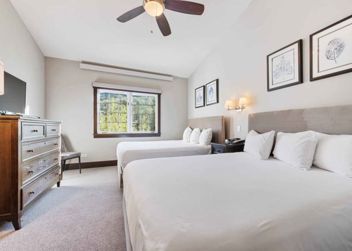 A spacious bedroom at The Charter B440 with two queen beds, each with white bedding and light beige headboards, a dark wood nightstand between them, and framed artwork on the walls, with a window offering a view of greenery.