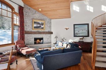 A cozy and elegant living room at Jay Chalet, featuring a large stone fireplace, comfortable blue sofas, and a stunning view of a snow-covered winter landscape through a grand arched window.
