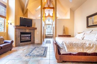 The luxurious bedroom at 213 County Road 1041 features a cozy fireplace, a comfortable bed, and an adjoining bathroom with a Jacuzzi tub, all under a soaring vaulted ceiling with exposed wooden beams.