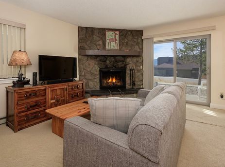A cozy living room in Meadow Ridge 18 featuring a fireplace, a comfortable sofa, a wooden TV console with a flat-screen TV, and large sliding glass doors leading to an outdoor view.