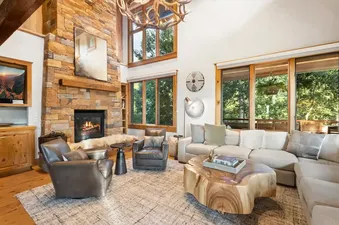 A spacious living room in Telemark A with a stone fireplace, light-colored sectional sofa, two dark leather armchairs, a live-edge wood coffee table, and large windows looking out to green trees.