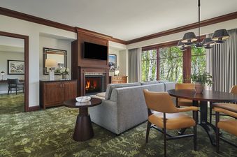A spacious and elegantly furnished living area inside the Montage Deer Valley Accessible Resort View King Suite, with a fireplace, comfortable seating, and a dining table, featuring large windows that offer a view of green trees outside.