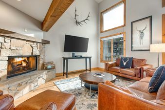 A rustic yet luxurious living room at Deer Valley 355 B, featuring a stone fireplace, leather sofas, a flat-screen TV, and large windows with wooden frames.