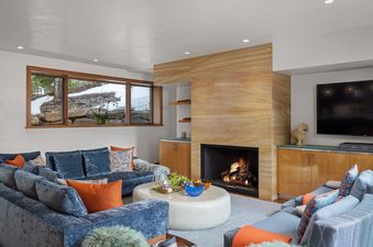 A cozy and modern living room features blue velvet sofas with orange pillows, a round white coffee table, and a sandstone fireplace, with a snowy outdoor view through large windows.