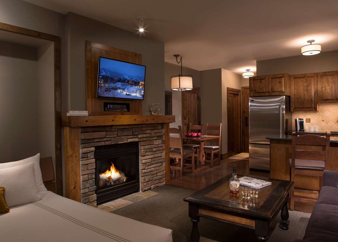 A cozy view of the Teton Mountain Lodge Alpine Studio interior featuring a stone fireplace, a wall-mounted TV, a partial bed, and a kitchen area.