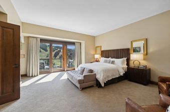A bedroom with a king-sized bed, a large window overlooking a mountain view, a plush ottoman, two armchairs, and a bedside table with a lamp.