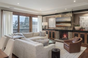 A spacious living room features a fireplace, a sectional sofa, a television, and a large window with an exterior view of snow-covered trees.