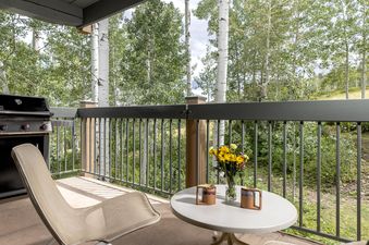 A private balcony featuring a BBQ grill, outdoor seating, and a table with fresh flowers, set against a backdrop of aspen trees.