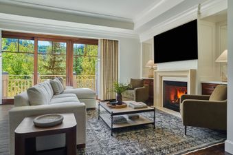 A spacious living room featuring a large sectional sofa, a fireplace with a mounted television, and a balcony with expansive views of lush green trees.
