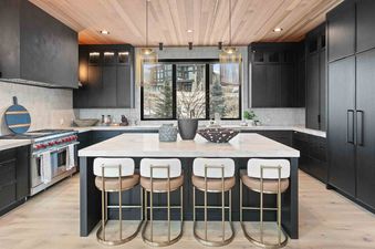 A modern kitchen with dark cabinetry, a white marble island with four bar stools, and a large window looking out onto a winter landscape.