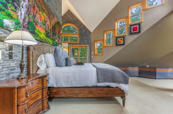 A uniquely designed bedroom at the Intrepid Retreat, featuring a bed with white linens, a wooden nightstand, a large nature artwork, and several wooden-framed windows offering exterior views.
