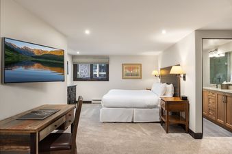 A hotel bedroom with a large bed, wooden furniture, a wall-mounted TV, and an adjacent bathroom.