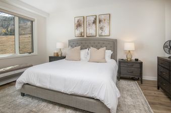 A cozy master bedroom features a king-size bed with white linens, two nightstands with lamps, abstract art, and a large window overlooking a mountain landscape.
