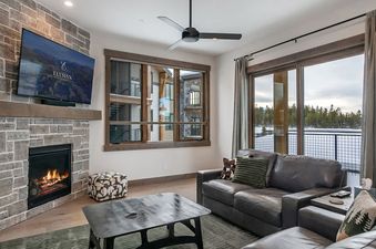 A cozy living room featuring a stone fireplace with a mounted flat-screen TV, a dark leather sofa, and large windows offering scenic outdoor views.