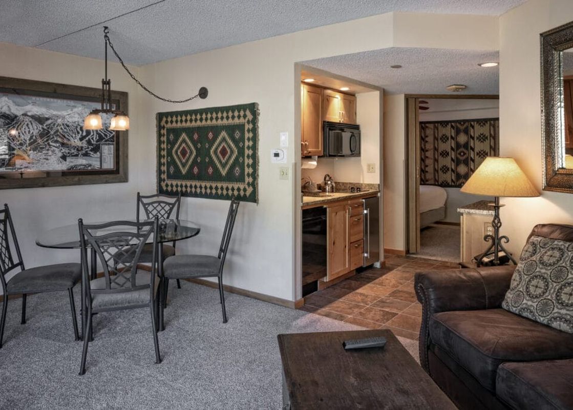 A view of the dining and living area inside the Colorado Suite Beaver Run Resort, featuring a glass dining table, leather sofa, and a compact kitchen.
