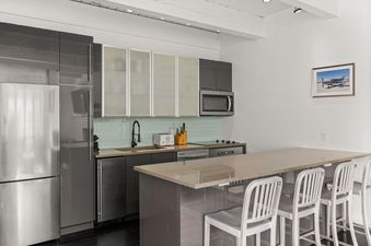 A modern kitchen with stainless steel appliances, grey cabinetry, a light blue subway tile backsplash, and a long kitchen island with four white bar stools.