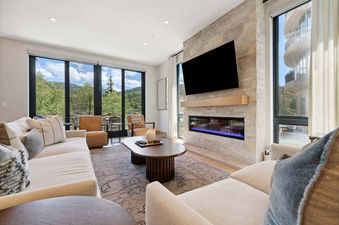 A modern and cozy living room with large windows overlooking green mountains, featuring a fireplace, flat-screen TV, sofas, and a coffee table.