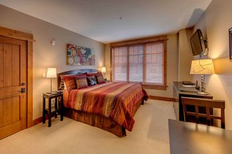 A bedroom with a king-sized bed, a large window with blinds, and a TV mounted on the wall.