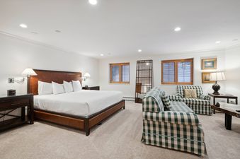 A spacious bedroom in The Charter L032 featuring a king-sized bed, a sitting area with gingham-patterned sofa and armchair, and large windows with wooden blinds.