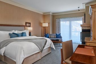 A spacious bedroom features a large bed with white linens and blue accent pillows, a sitting area with a sofa, and a desk, all illuminated by natural light from a large window.
