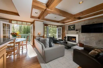 A spacious and elegant living room with a stone fireplace, flat-screen TV, comfortable seating, and French doors opening to a balcony with mountain views.